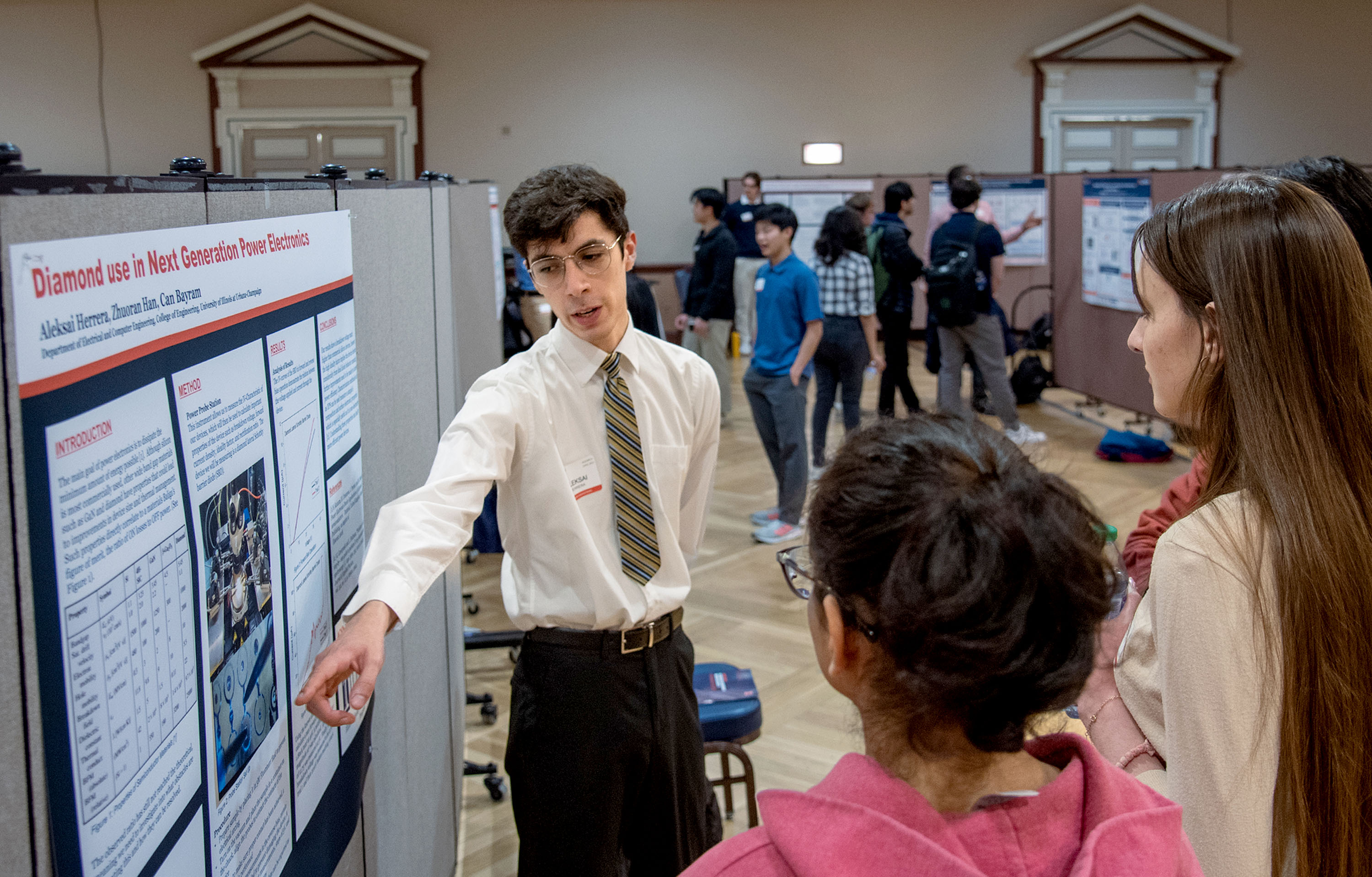 A professionally dressed student points at a research poster explaining his work to an audience of other undergraduate students in the Illlini Union Ballroom.