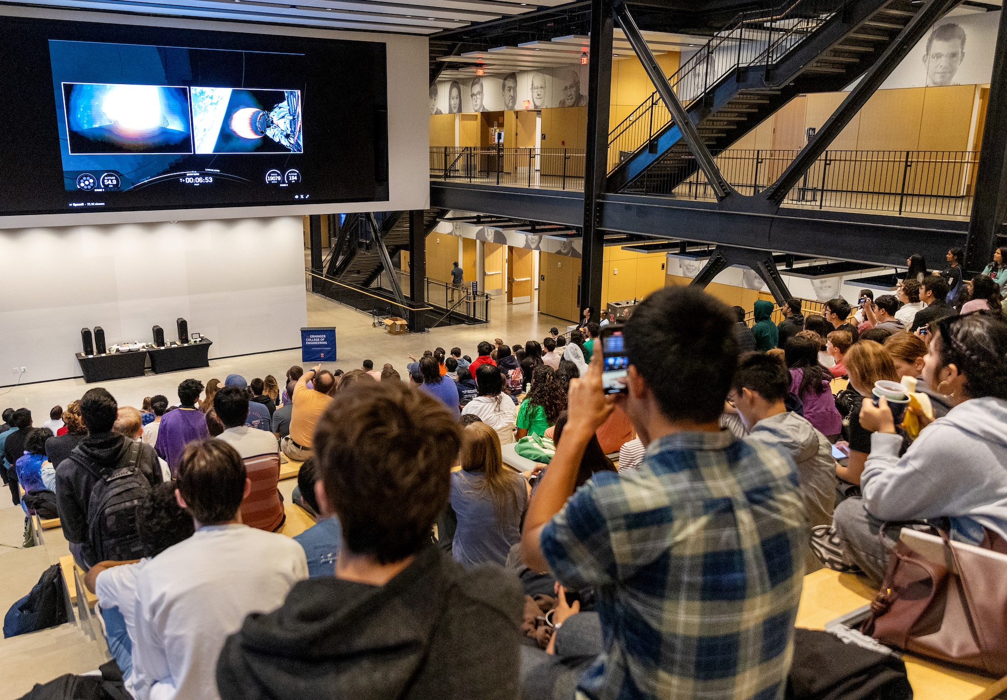 A view of the Carruthers Geocorona Observatory launching into space on a large screen in front of a large group of students and community members.