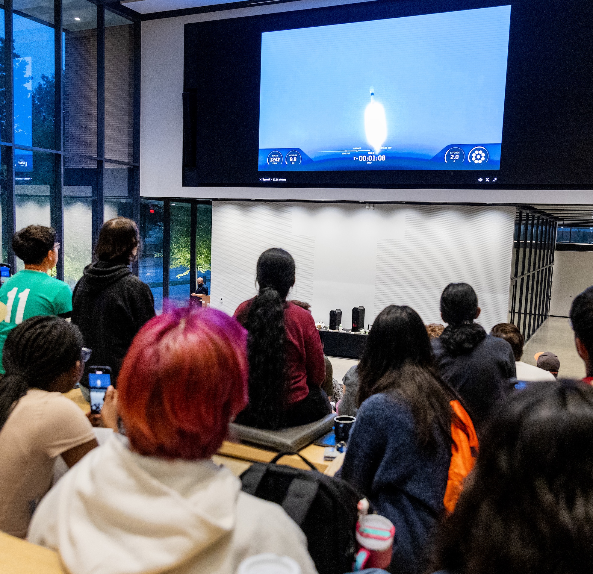 A view of the Carruthers Geocorona Observatory launching into space on a large screen in front of a large group of students and community members.