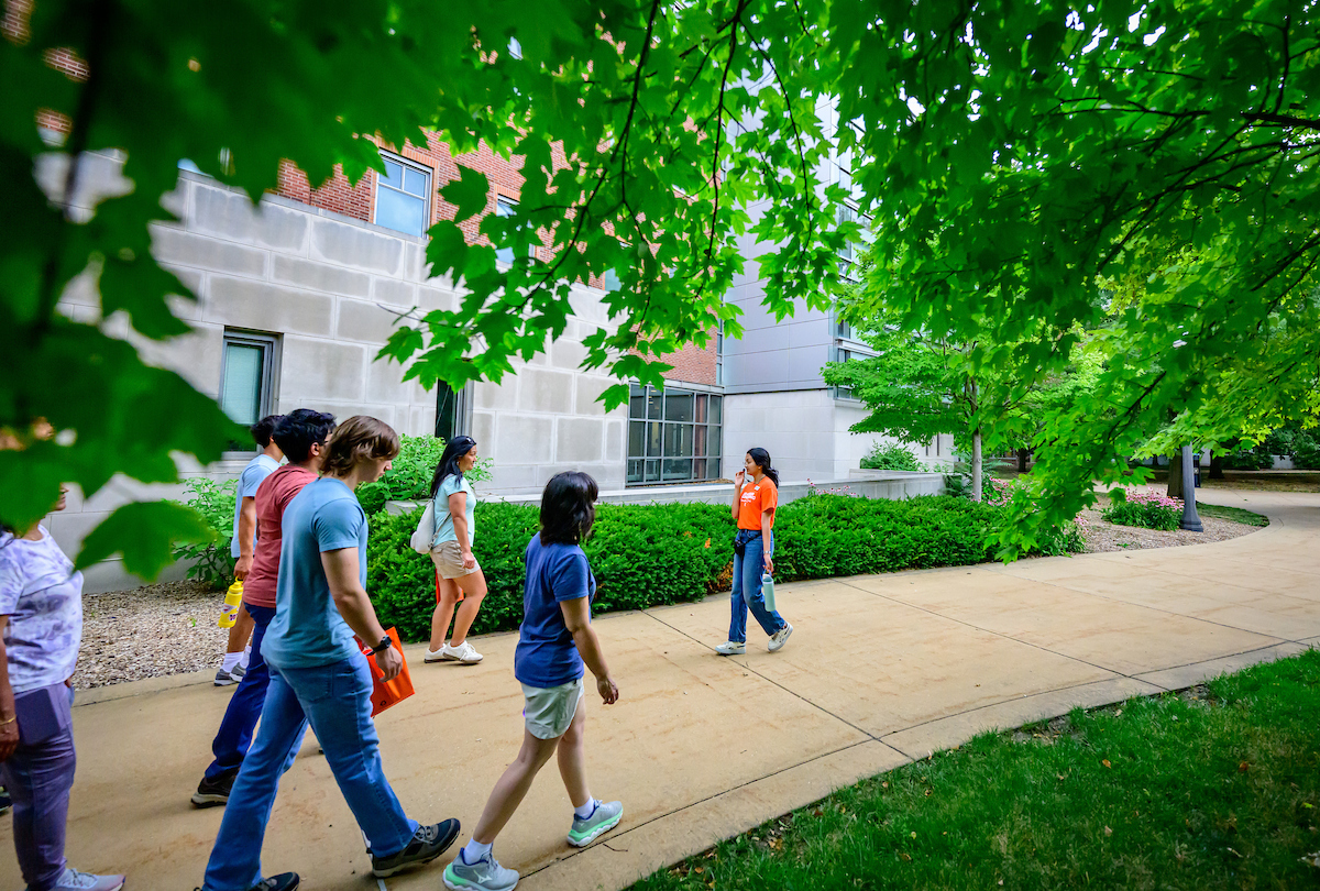 A Grainger Engineering tour guide walks backward, talking to a group of visiting students and families, in front of the Thomas M. Siebel Center for Computer Science.