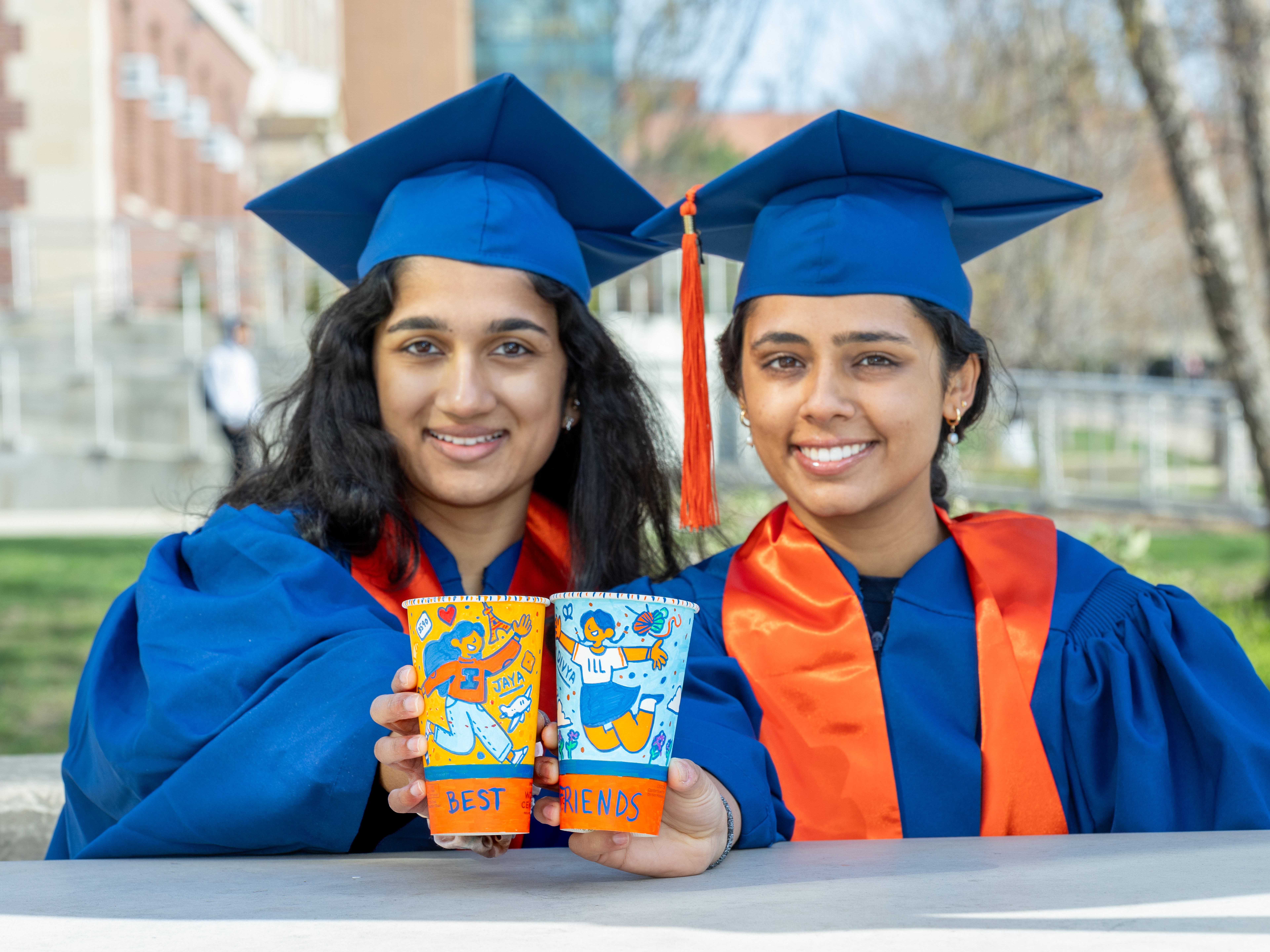 Divya and Jaya in their graduation gowns, holding their colorful Illinois &amp;quot;Best Friends&amp;quot; cups on the quad.