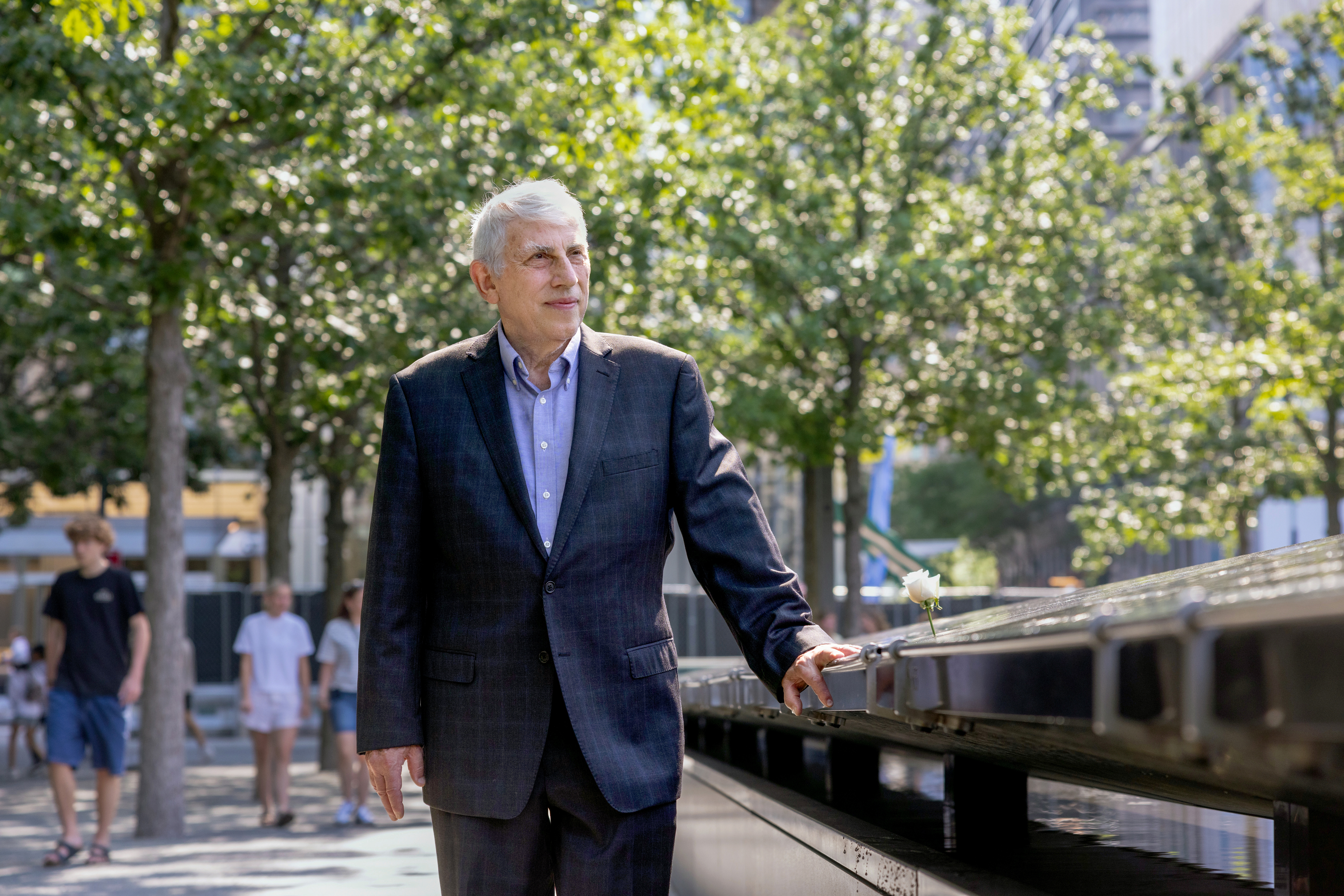 Bob Smilowitz walking along the sidewalk adjacent to the September 11 Memorial in New York City, with trees in the background.