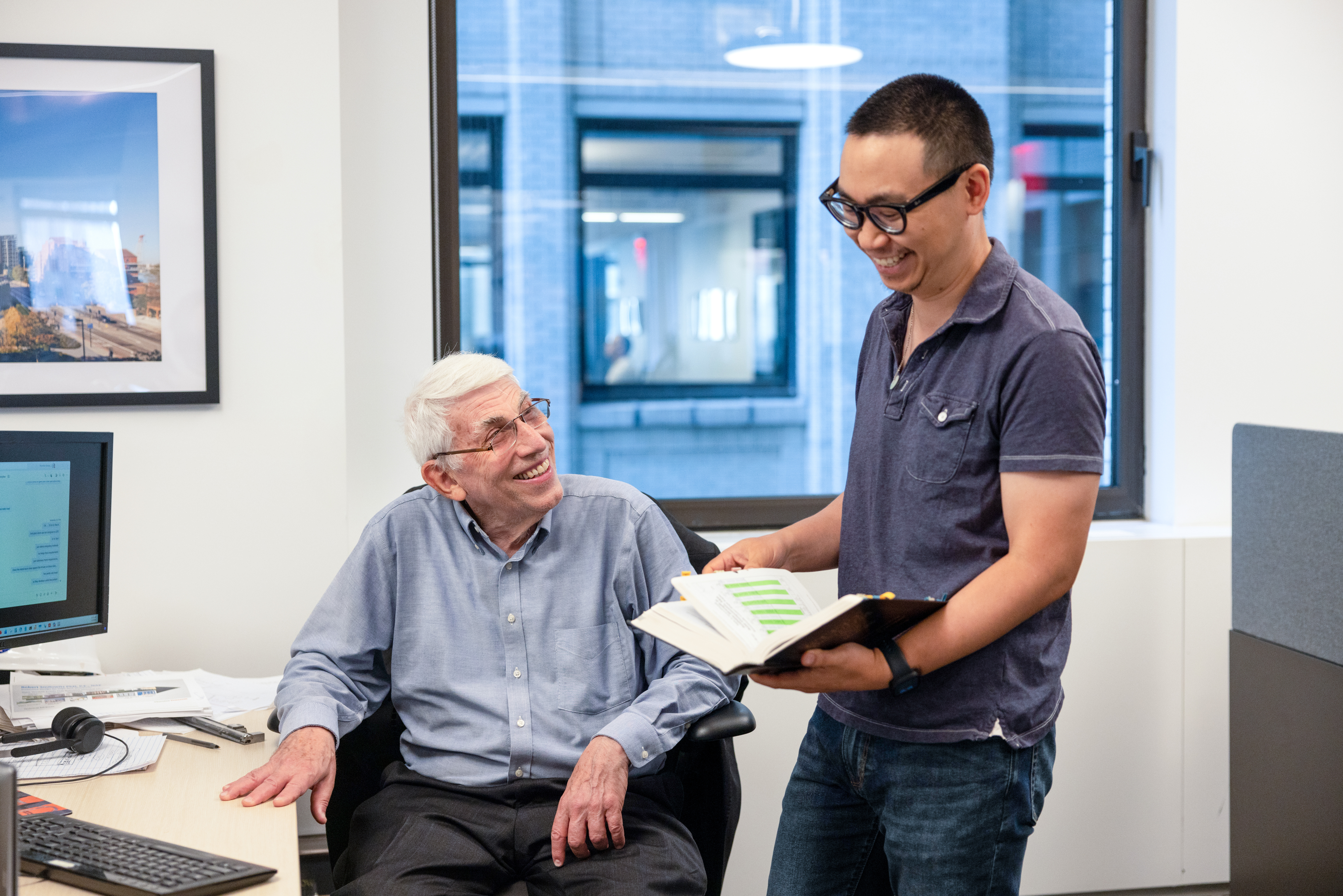 Bob Smilowitz (sitting) and a colleague (standing) smile as they review a file of papers inside Bob's office.