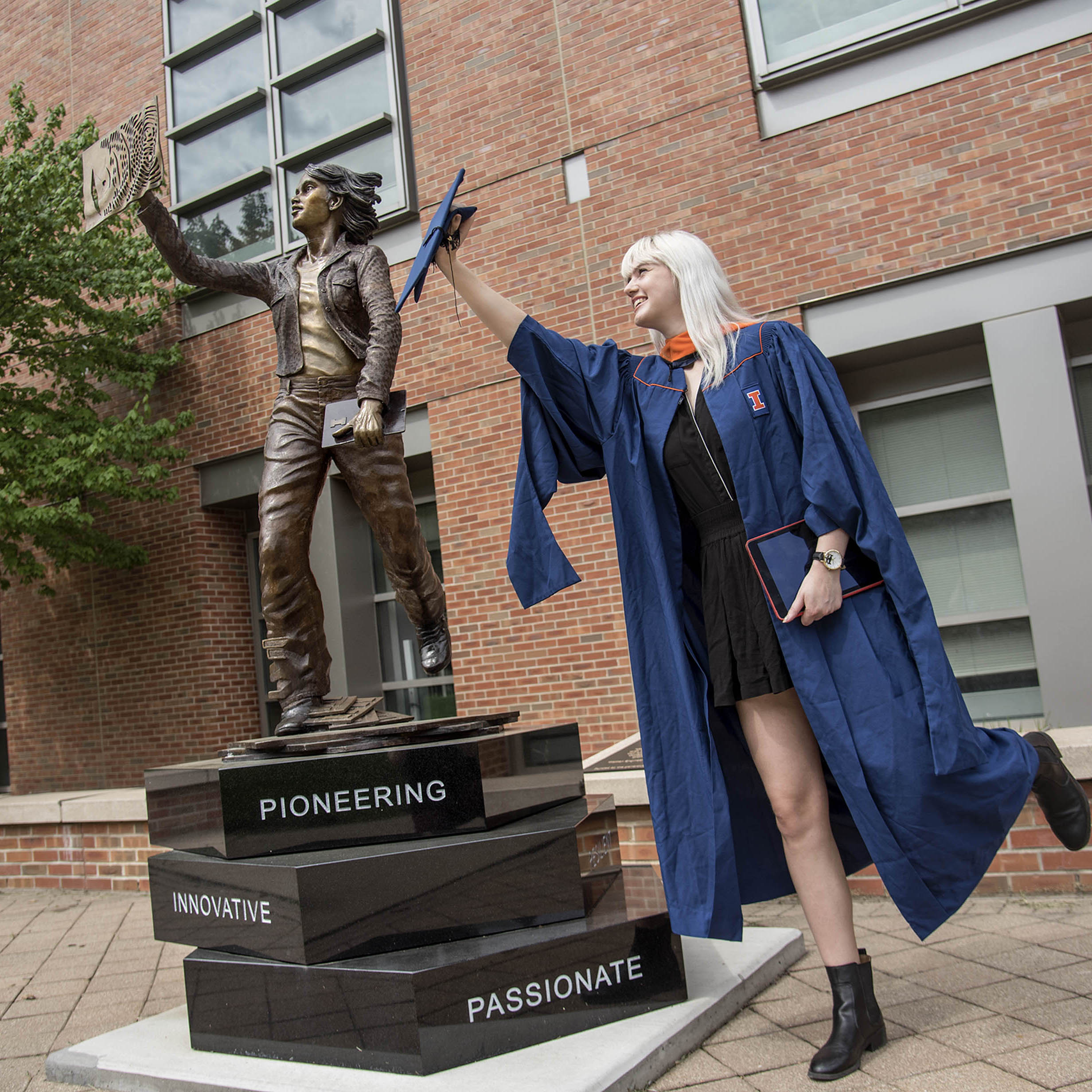 A blonde woman in her graduation regalia holding her graduation cap mimics the statue of 'The Quintessential Engineer'. The statue is of a woman looking up with one arm raised, a book in her other hand, and her back leg raised. She is standing on three large blocks, each with a word (pioneering, innovative, and passionate) engraved on it. 
