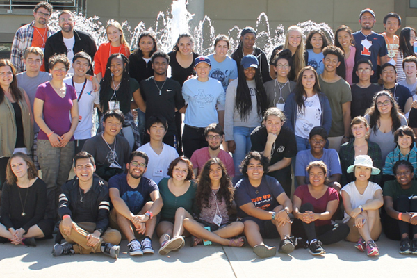 A large multicultural group of people standing together with their arms around one another in front of a fountain. 