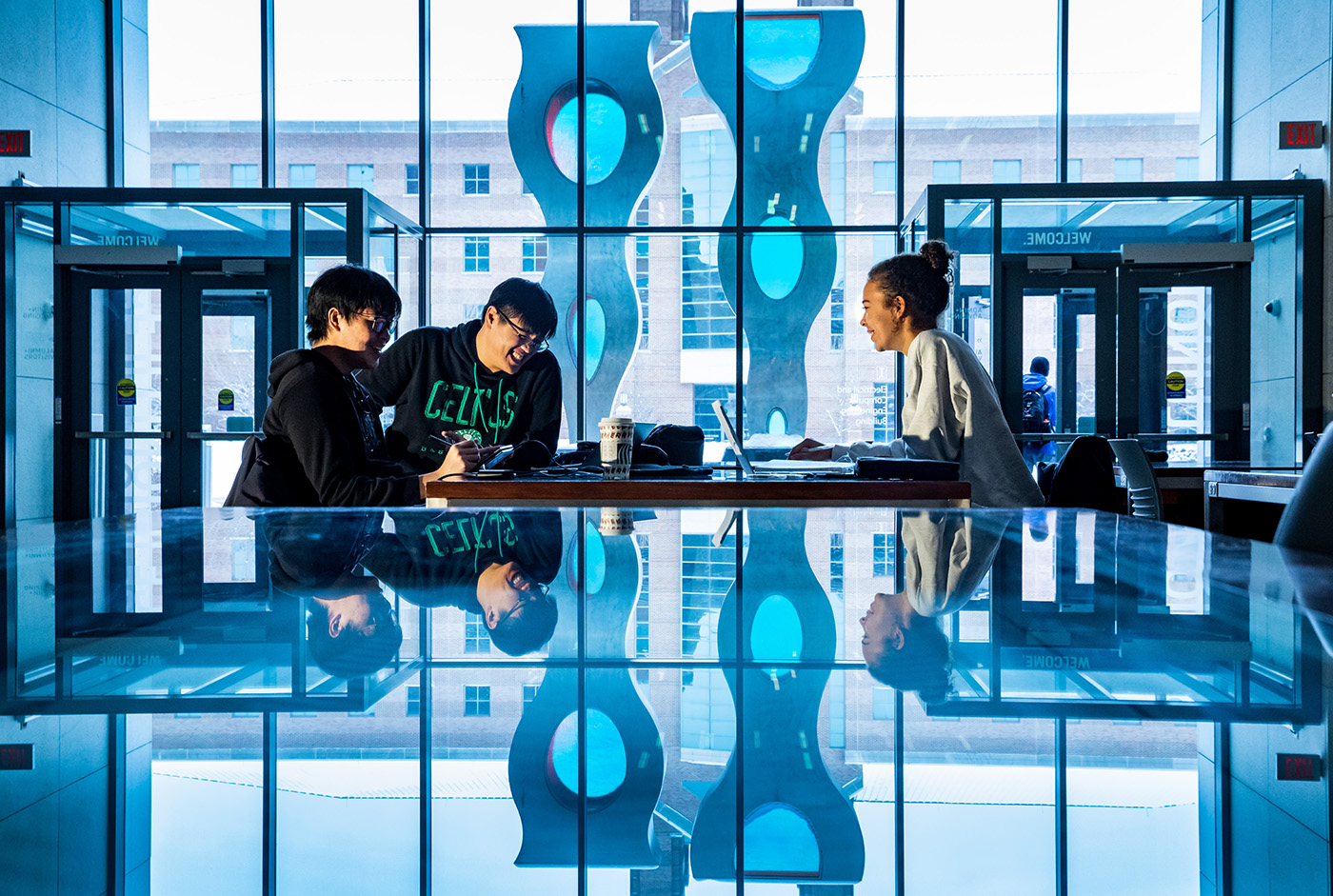 Three students sit around a table studying at the Electrical and Computational Engineering Building with windows onto the quad in the background. 