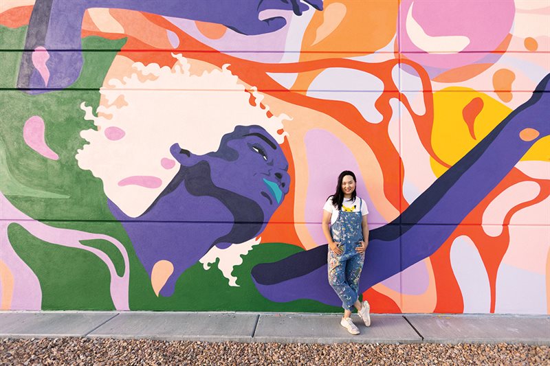 Amanda standing in front of the mural in paint splattered overalls