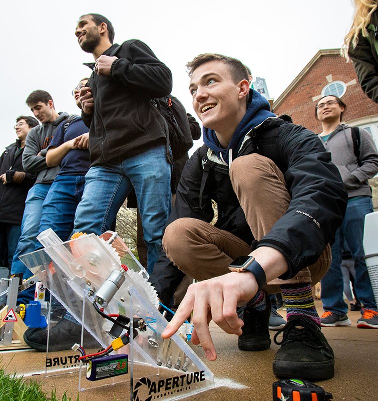 Student smiling as he operates a mechanical device. More students watch from behind