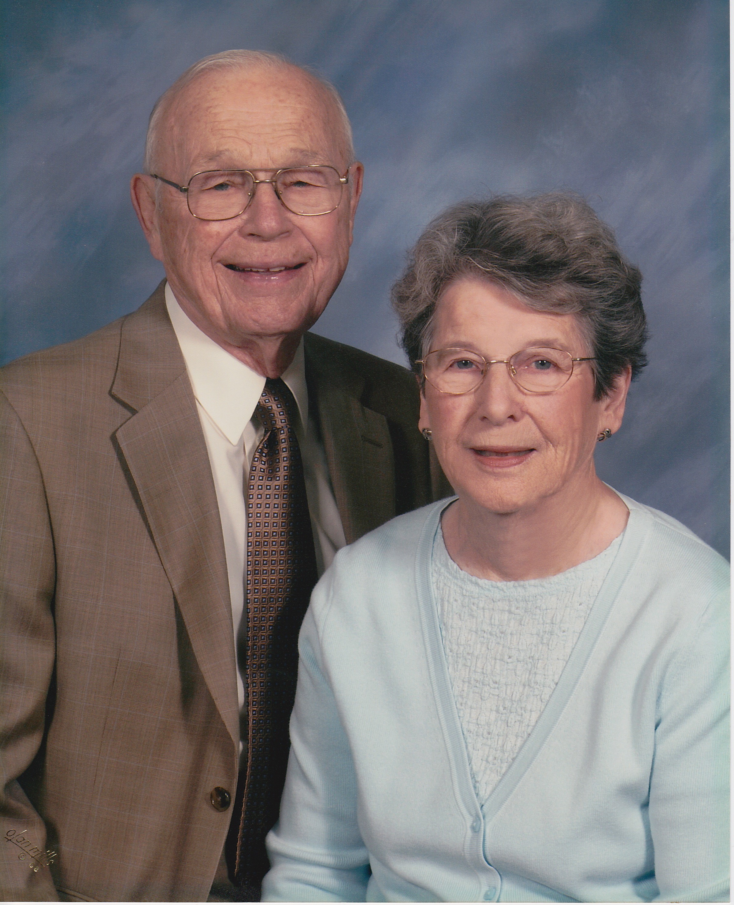 An elderly couple posing together in front of a gray back drop. A white man in a brown suit stands up with one shoulder tucked behind his white wife, who wears a light blue sweater. 