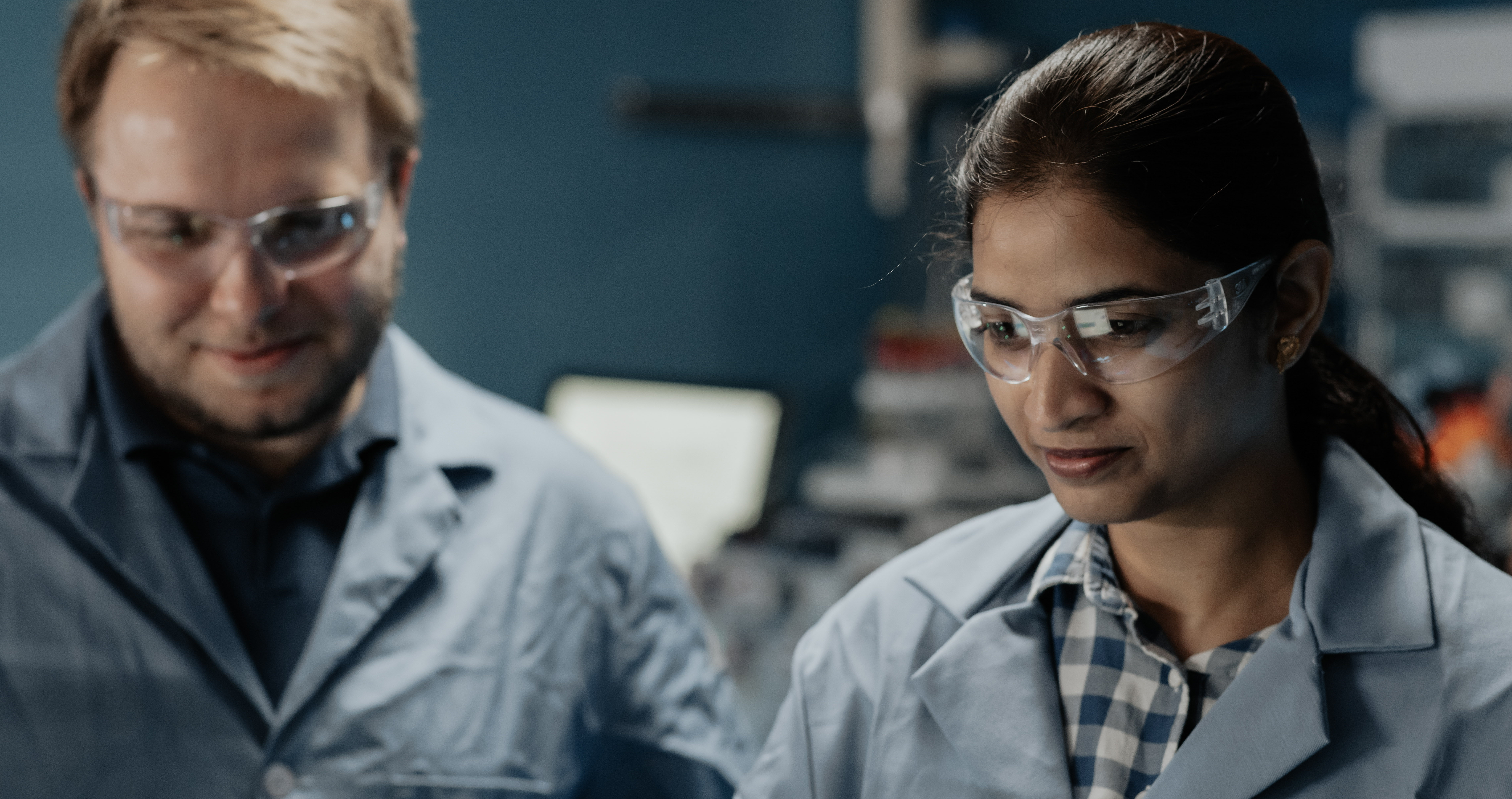 A man guides a woman in her research in a lab as they look at a screen with data. They both wear blue lab coats and safety glasses. 