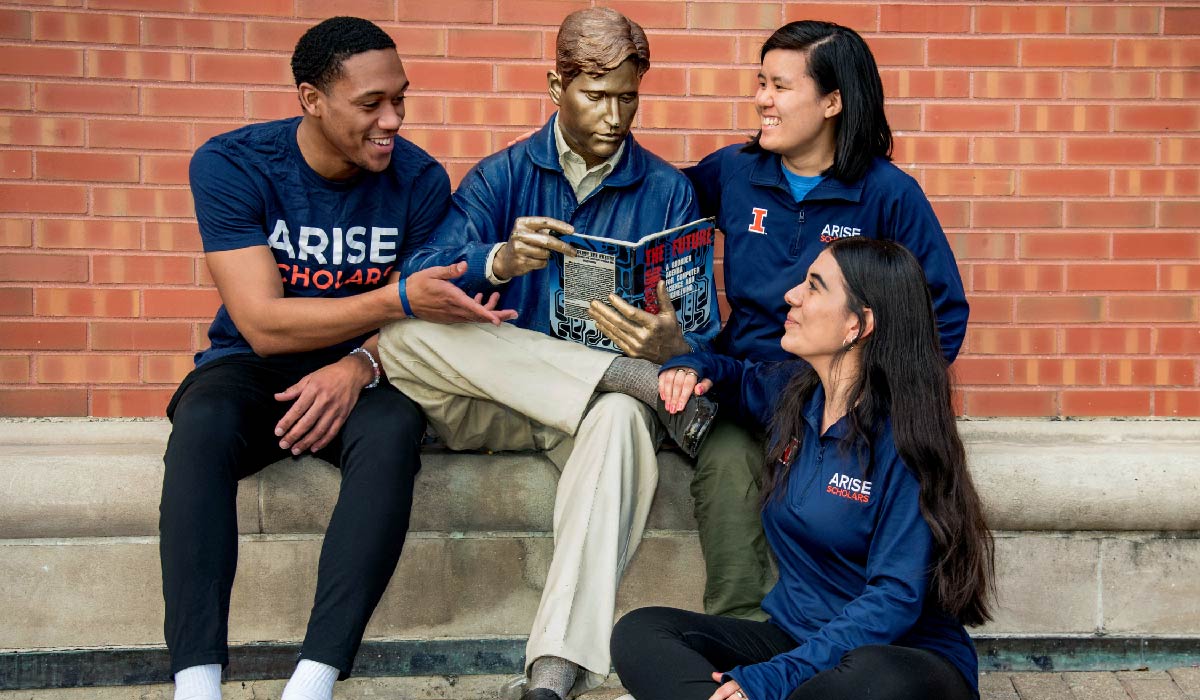 Three ARISE undergraduate students in matching 'ARISE' shirts sit around Grainger Bob, a metal statue of a life-size man sitting and reading a textbook, outside of the Grainger Library. 