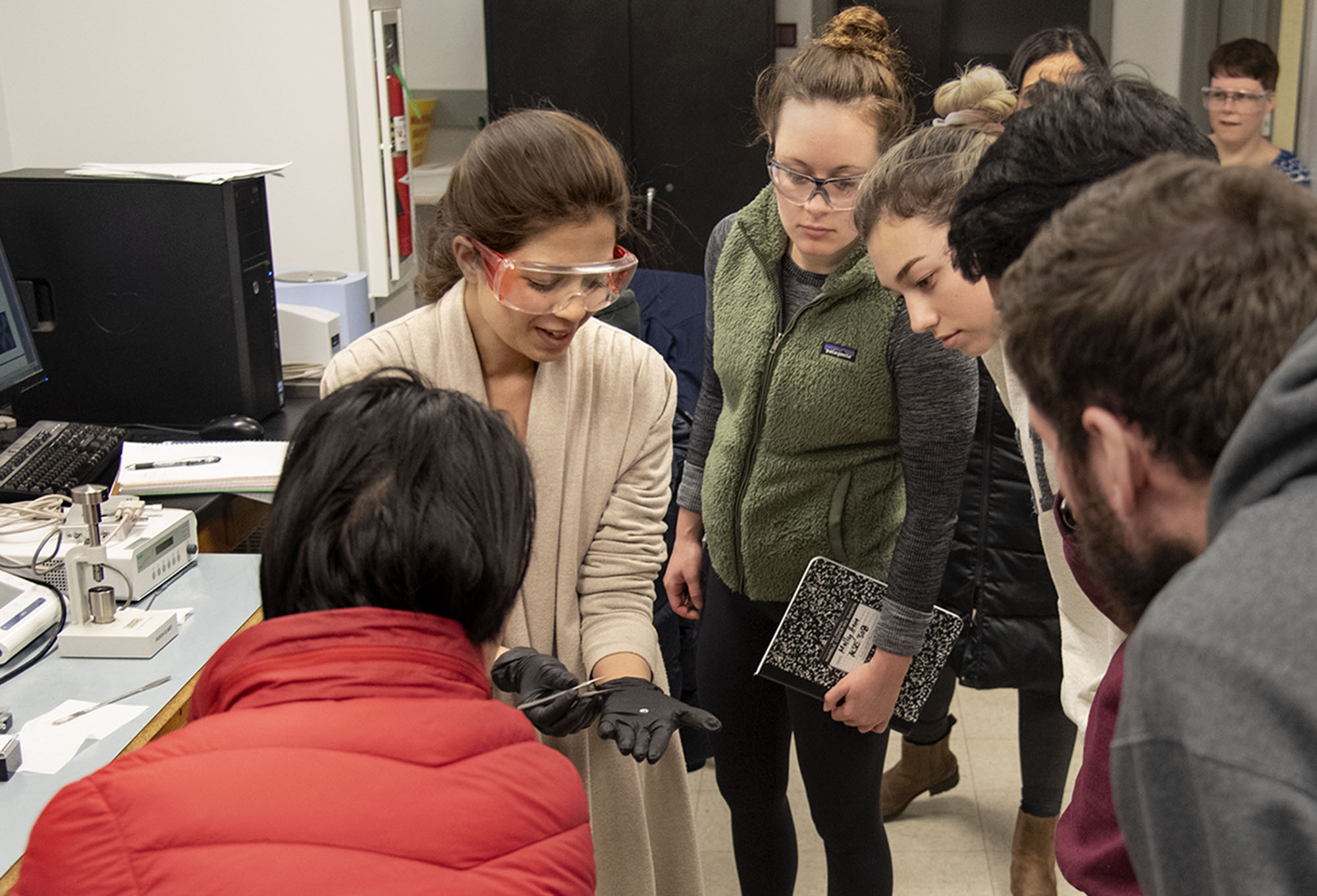 A  teaching assistant demonstrates how to use a Differential Scanning Calorimetry in MSE 308 lab to a group of students gathered around her.