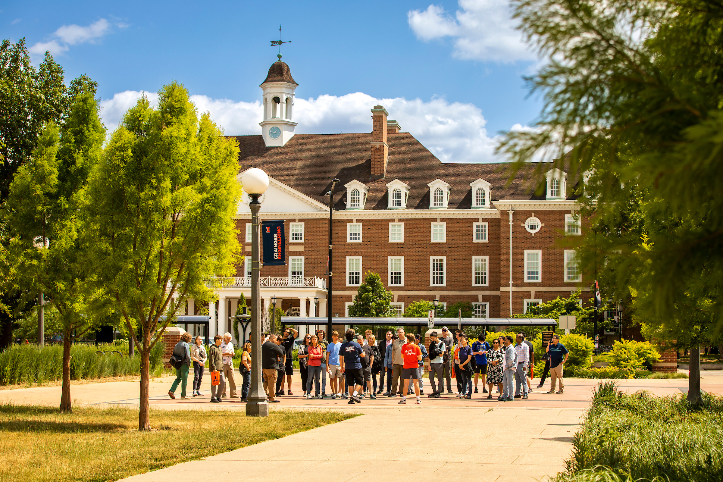 A group of people led by an undergraduate tour guide meet in front of the Illini Union, a tall white and brick building. 