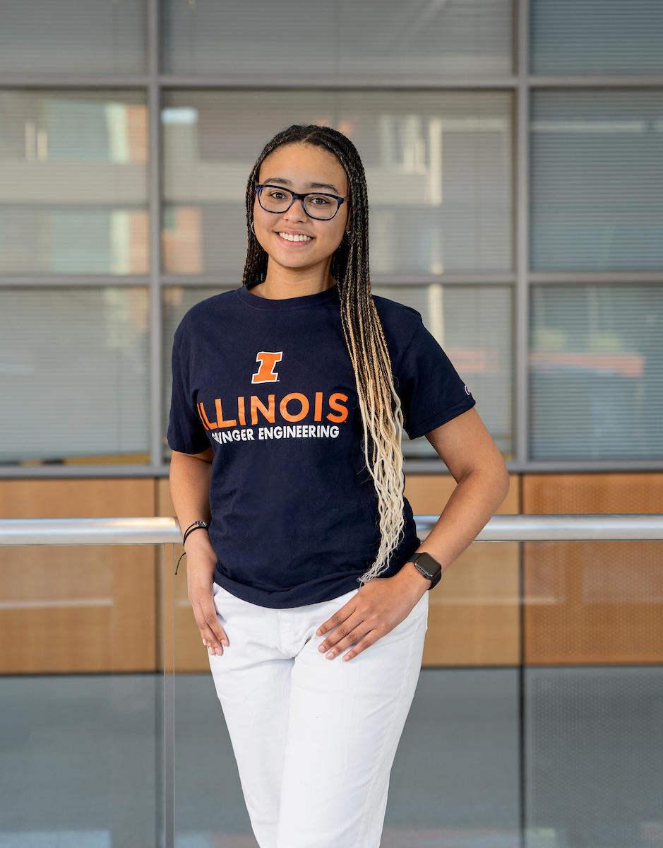 A woman stands posing with her arms on her hips looking at the camera. She has hip-length ombre braids from brown to blond, and glasses. She wears a navy t-shirt that says "Illinois Grainger Engineering" and white pants. 