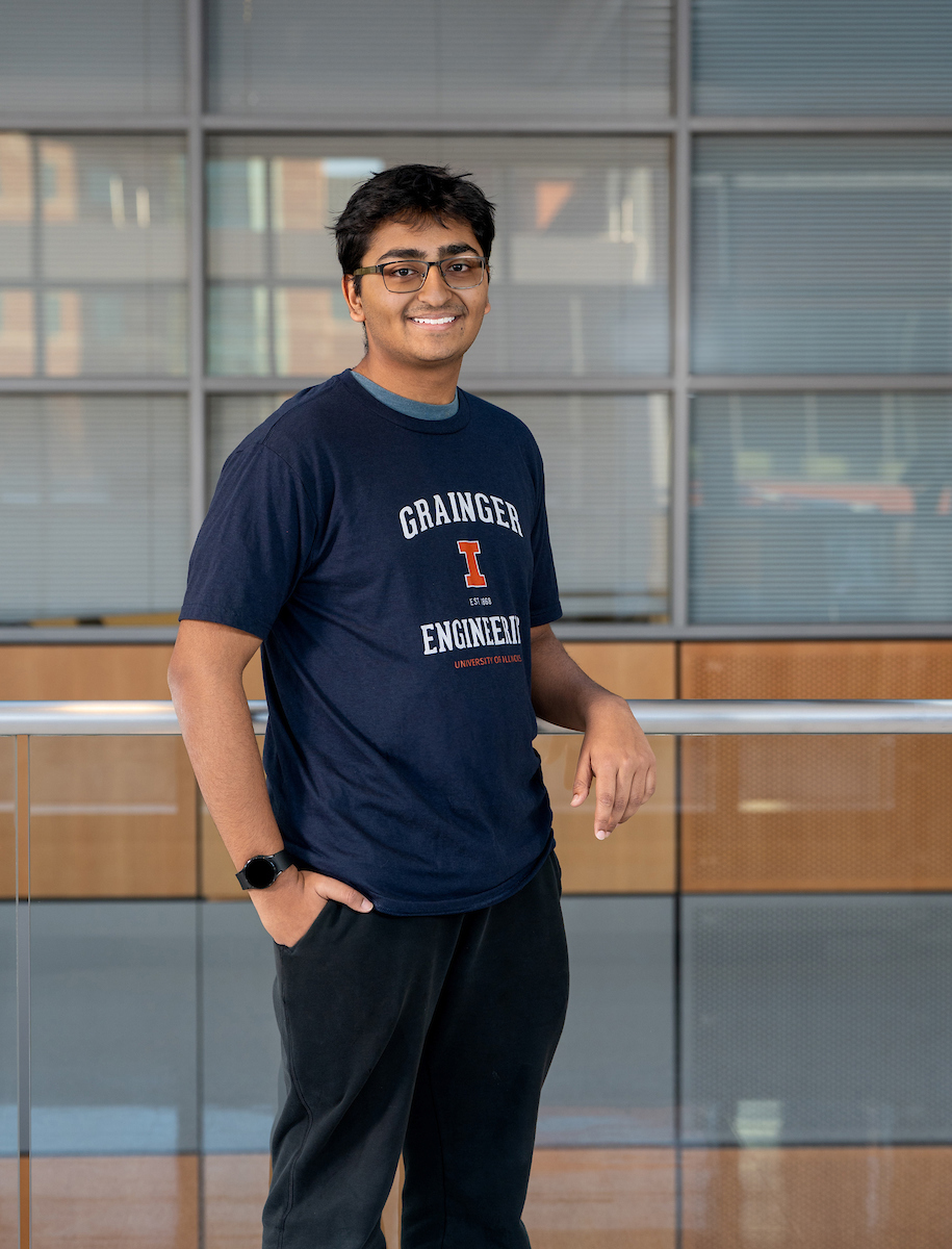 A young man stands posing for the photograph with one arm on a railing behind him and the other in his pocket. He had glasses and it wearing a "Grainger Engineering" shirt and black pants. 