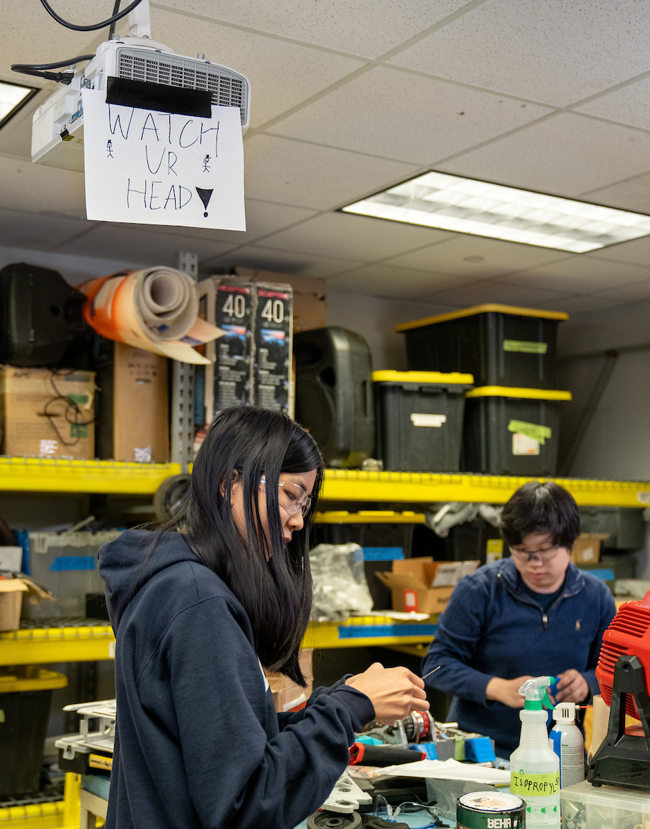 A female and male student on the iRobotics team work in the Engineering Student Projects Laboratory, with storage in the background.