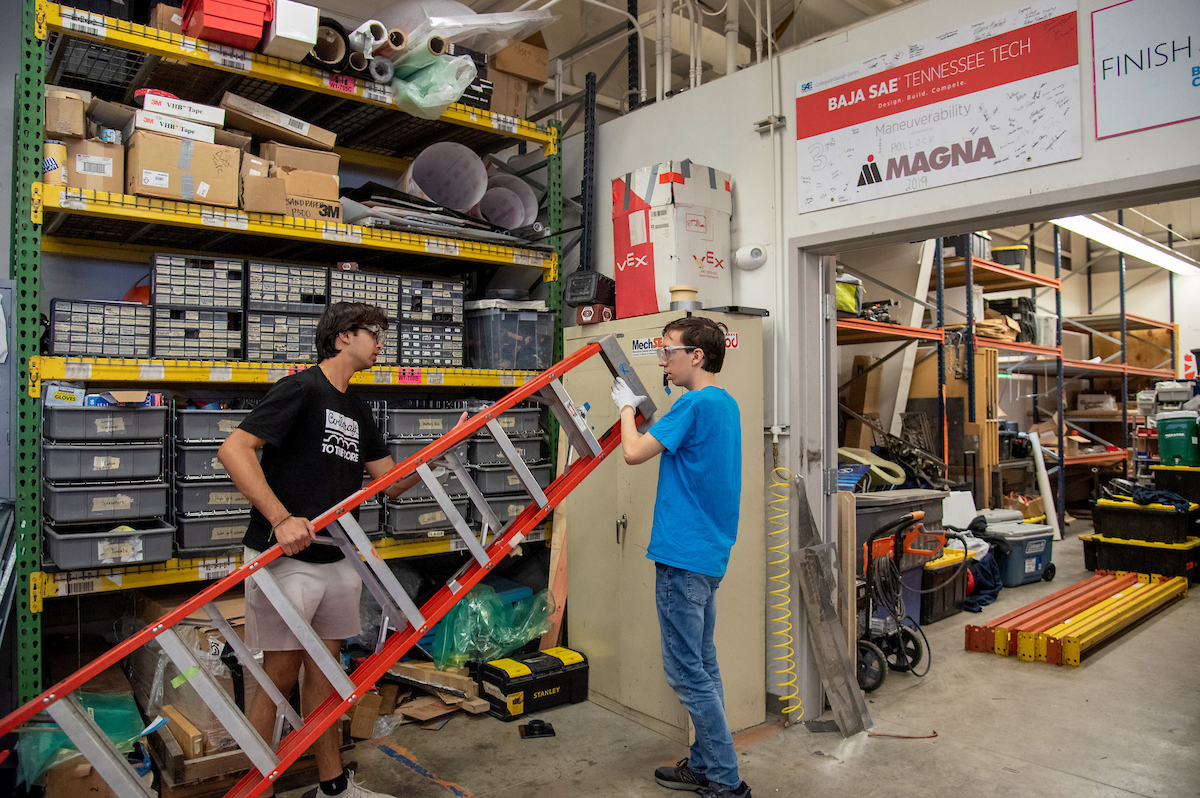Two male students moving a large ladder through a storage area.