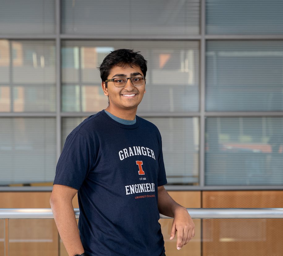 A young man stands posing for the photograph with one arm on a railing behind him and the other in his pocket. He had glasses and it wearing a &amp;amp;quot;Grainger Engineering&amp;amp;quot; shirt and black pants.