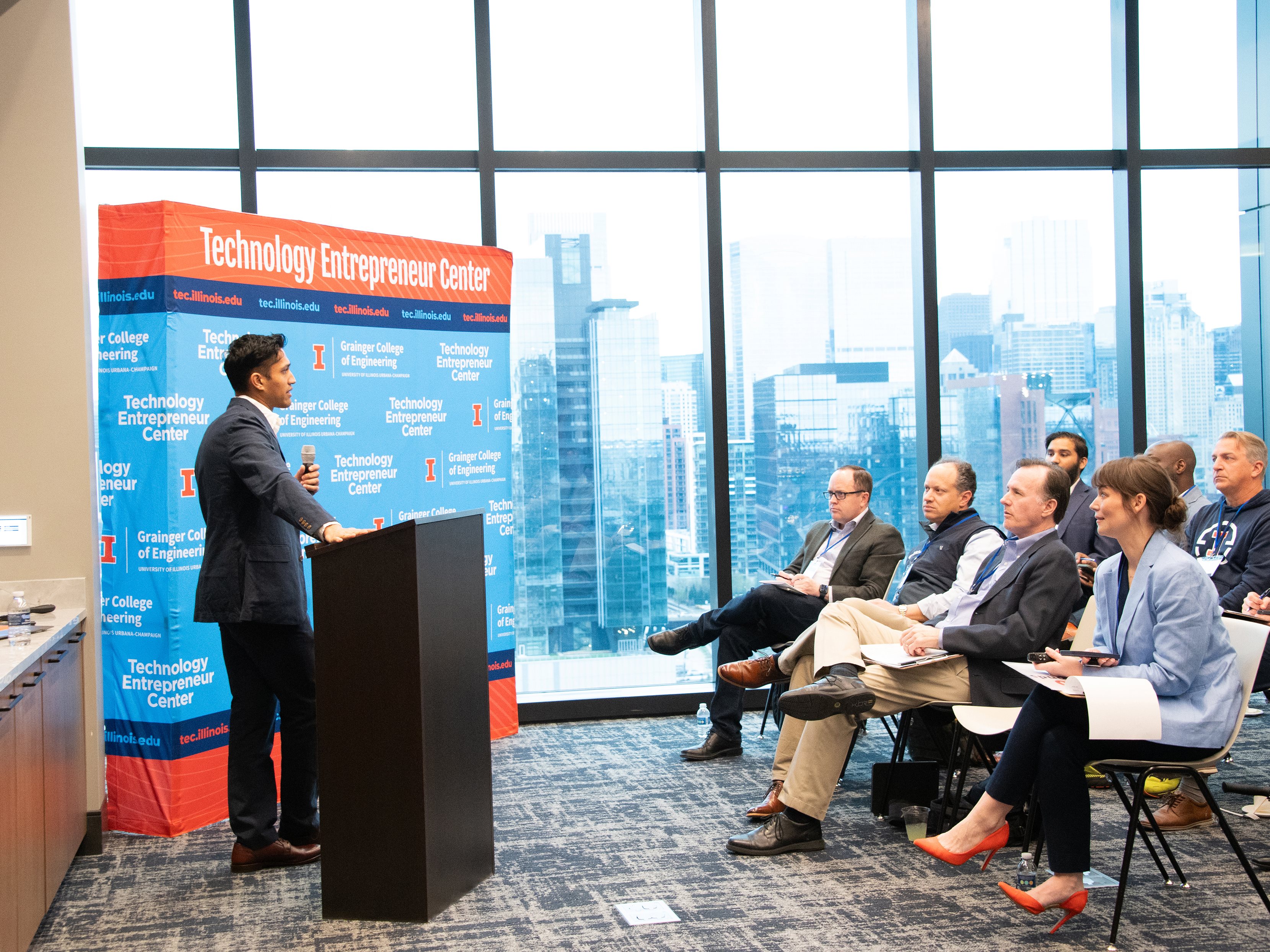 A male student, wearing a suit and standing behind a podium, pitches a start-up idea to an audience with the Chicago skyline in the background.